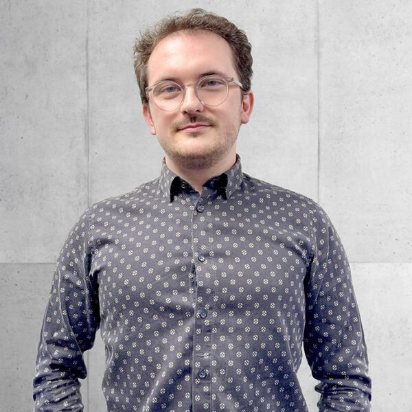 Benjamin Duhigg, a Senior Structural Technician at Peter Dann Ltd, wearing a shirt in front of a cement wall at Peter Dann