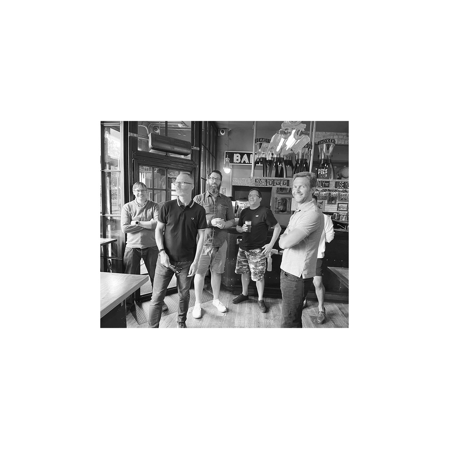 Five men posing in a bar like a boy band, one of them holding darts, in moody monochrome