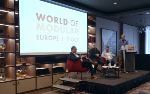 Inside a cosy conference on modular building with three men discussing the delights of modular construction in front of a screen that reads 'WORLD of MODULAR 2024'