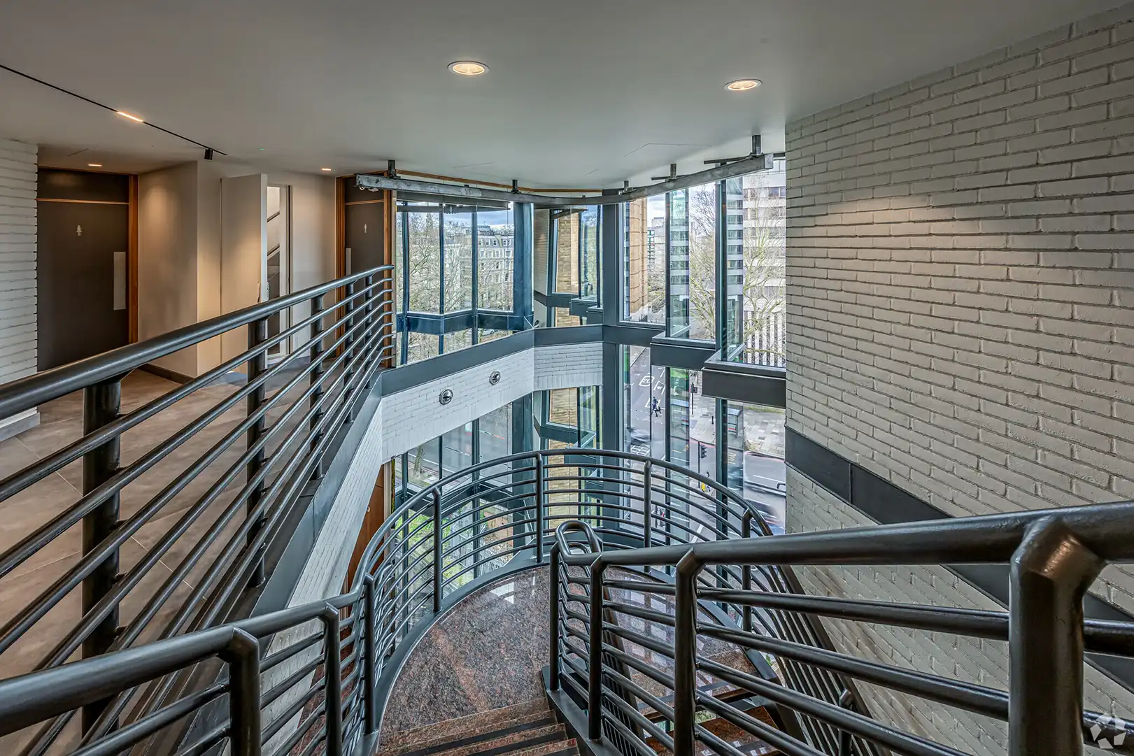 Contemporary spiral stairway in an office building with pale brick wall to the right and concertina glass facade ahead, with a landing to the left and grey wood doors